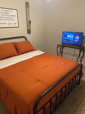 Uma cama com colcha laranja, luminária na parede e uma televisão sobre um suporte exibindo diversos canais.