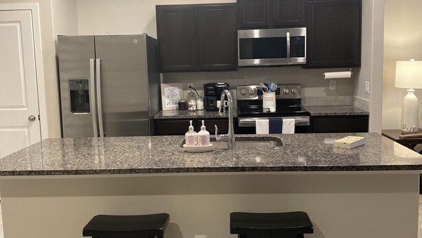 A kitchen with dark cabinets, stainless steel appliances, a granite countertop, and two bar stools. - Family Fun Meets Cozy Comfort - 3 Bedrooms, 2 Bathrooms, Game Room and Hot Tub! (Odessa)
