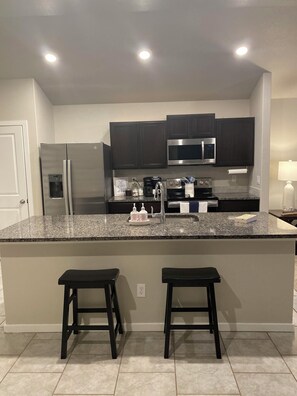 A kitchen with dark cabinets, stainless steel appliances, a granite countertop, and two bar stools. - Family Fun Meets Cozy Comfort - 3 Bedrooms, 2 Bathrooms, Game Room and Hot Tub! (Odessa)