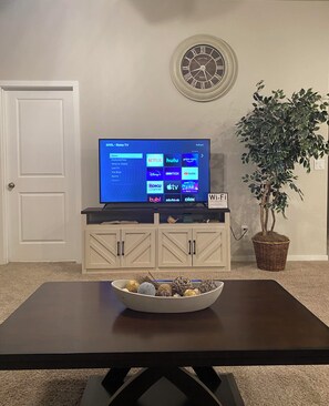 Een woonkamer met een TV via Roku TV, een klok aan de muur, een plant en een Wi-Fi signaal.