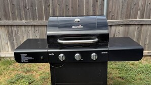 A black Char-Broil barbecue grill with three control knobs and a side burner, placed on a concrete pad in a backyard with a wooden fence in the background. - Family Fun Meets Cozy Comfort - 3 Bedrooms, 2 Bathrooms, Game Room and Hot Tub! (Odessa)