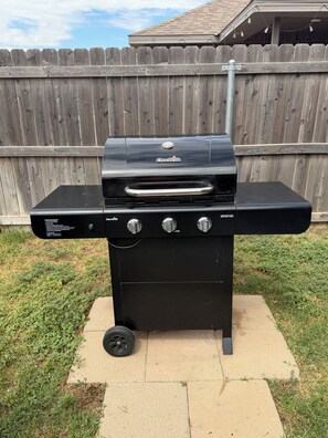 A black Char-Broil barbecue grill with three control knobs and a side burner, placed on a concrete pad in a backyard with a wooden fence in the background. - Family Fun Meets Cozy Comfort - 3 Bedrooms, 2 Bathrooms, Game Room and Hot Tub! (Odessa)