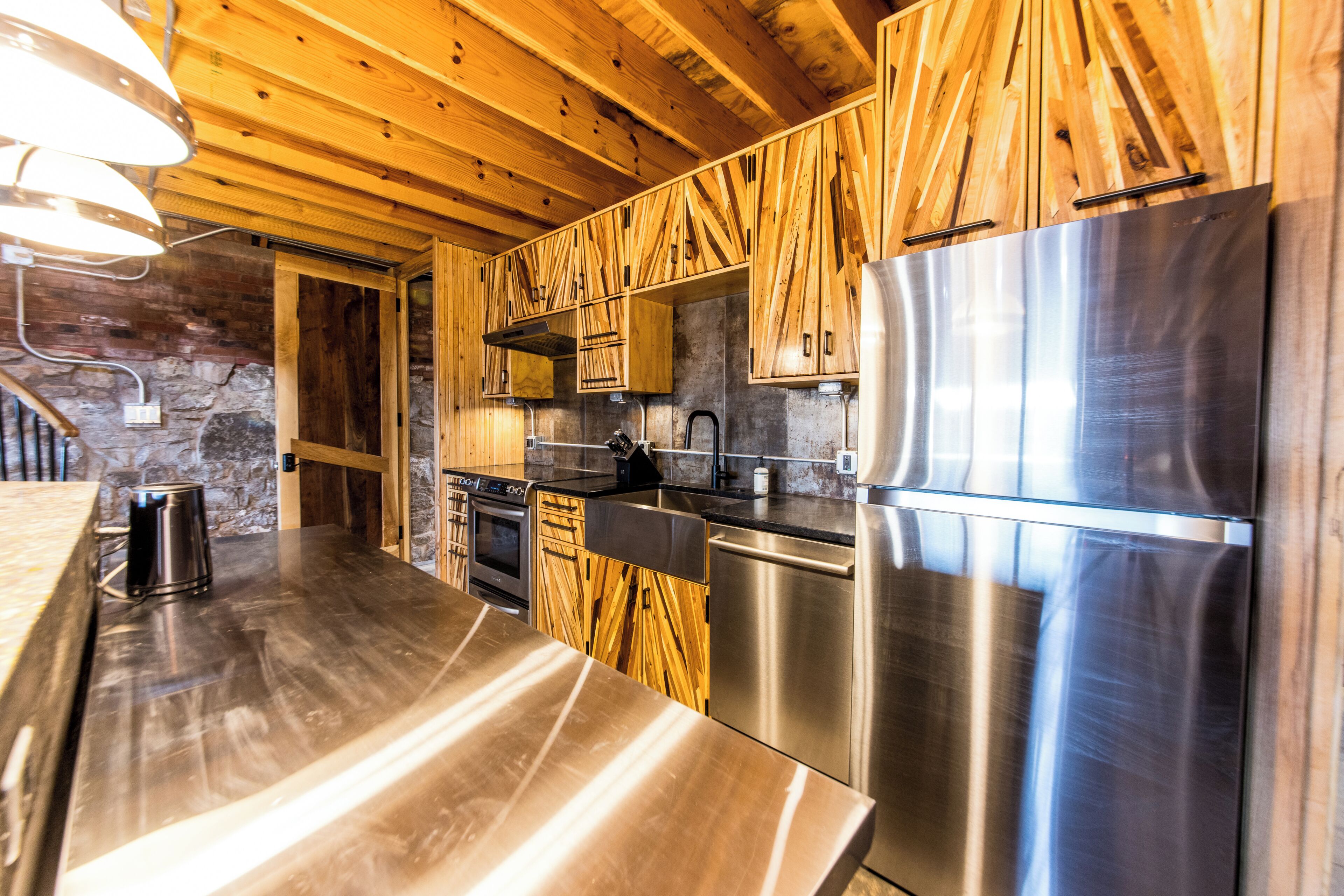 A modern kitchen with wooden cabinets, stainless steel appliances, and a stone wall.