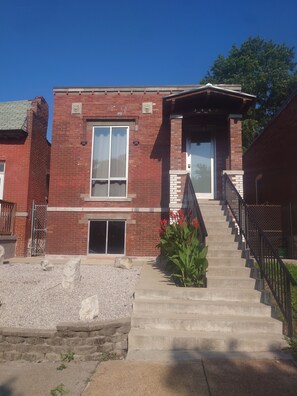 A two-story brick building with a front entrance, a staircase, and a small garden with red flowers. - The Lions Lair - New loft style home in STL (St. Louis)