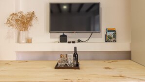 Un televisor de pantalla plana montado en una pared blanca, una mesita con una botella y vasos, un jarrón con flores secas y un libro titulado 'Portugal' en la pared.