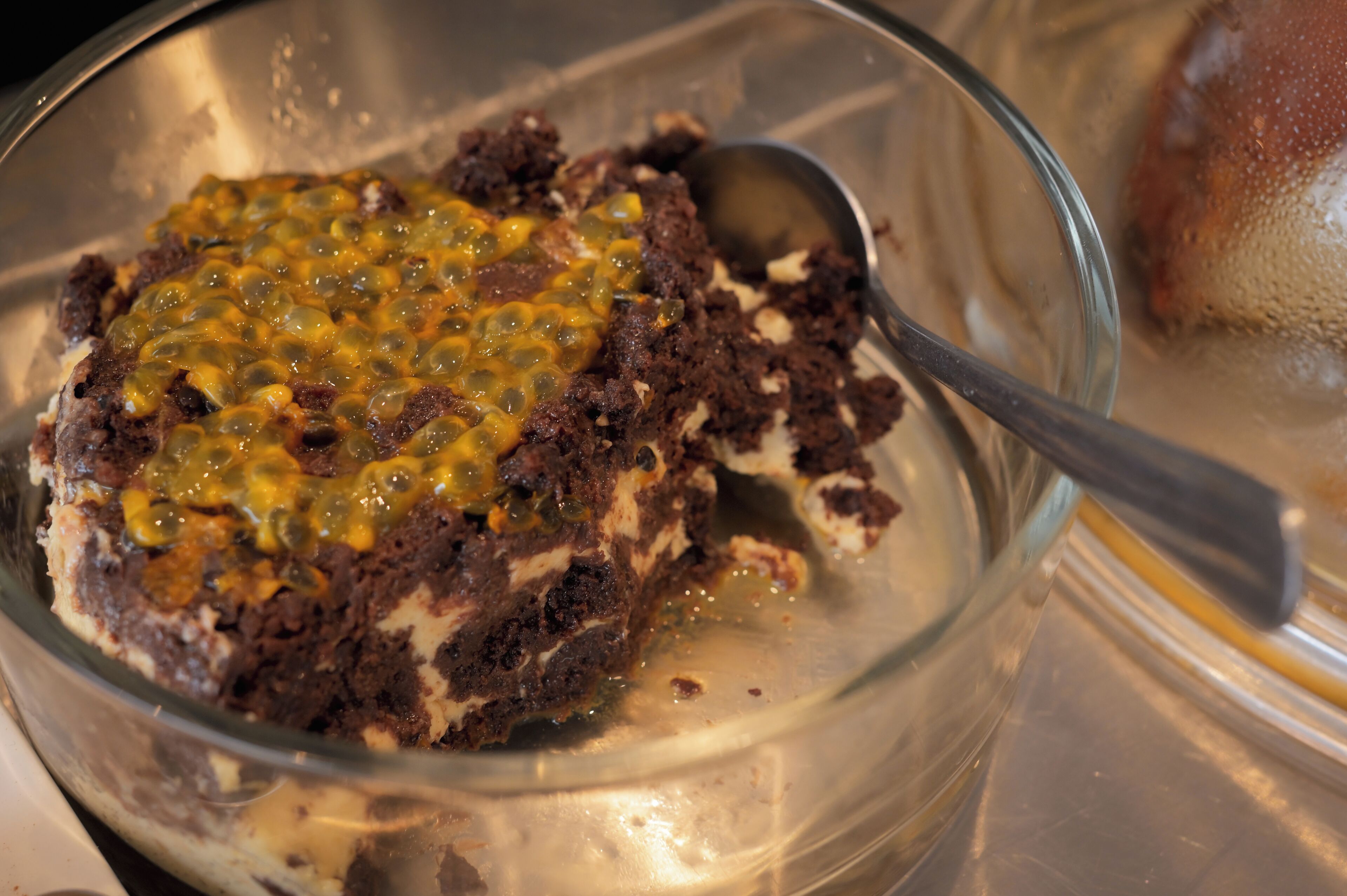 A dessert in a glass bowl with a spoon, containing a chocolate base topped with a yellow sauce and green toppings.