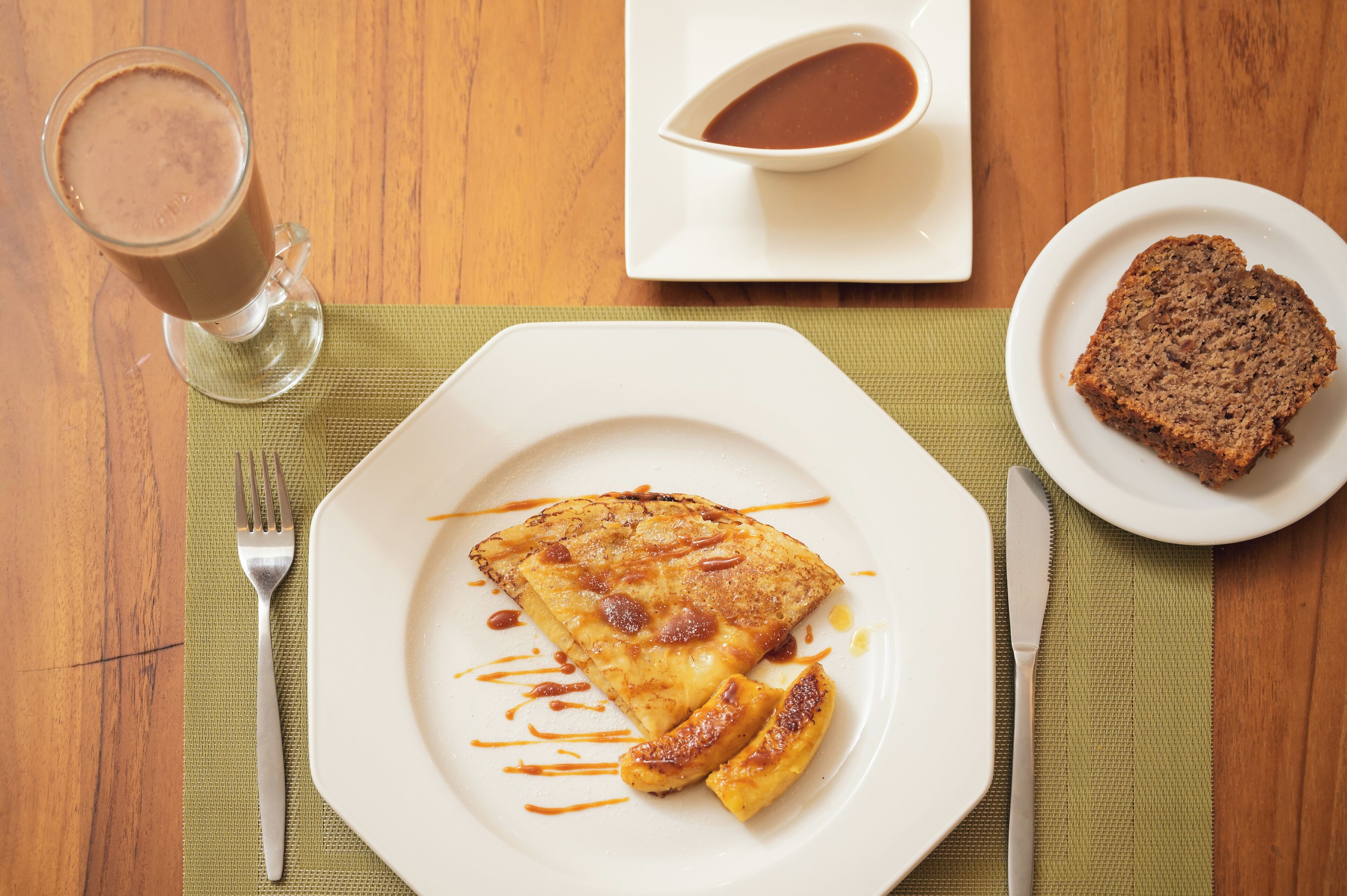 A breakfast setting with a plate of pancakes, a slice of bread, a cup of coffee, and a small bowl of sauce on a wooden table.
