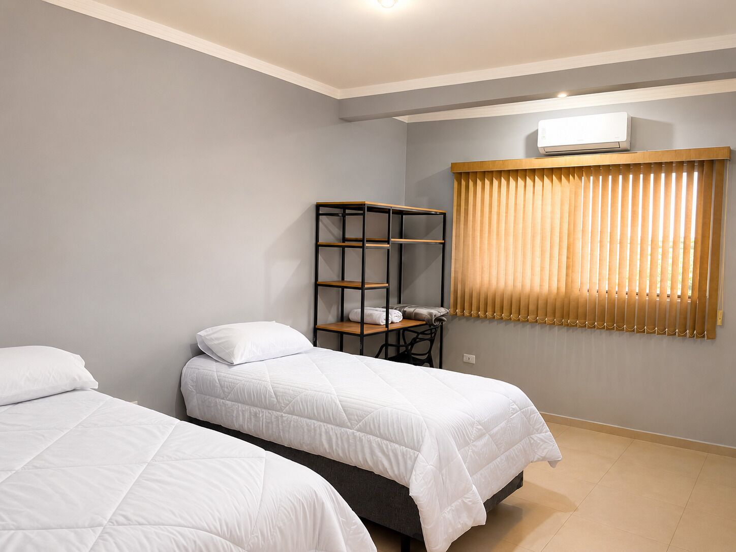 Habitación de hotel con dos camas, un escritorio, una silla, una ventana con persianas y un equipo de aire acondicionado.