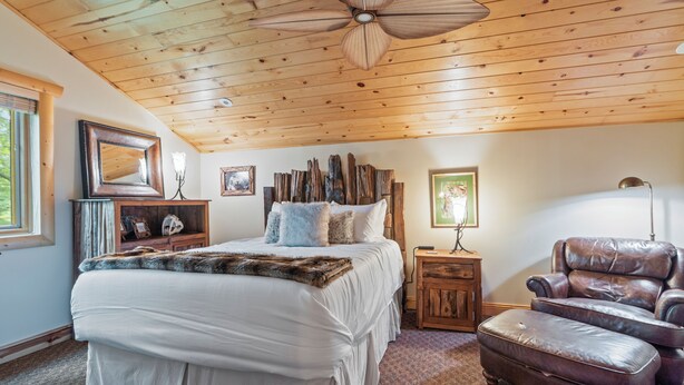 A bedroom with a wooden ceiling, a bed with white bedding, a leather armchair, and a nightstand with a lamp. - Handcrafted 4BR Cabin on Rabbit Lake, Aitkin (Aitkin)