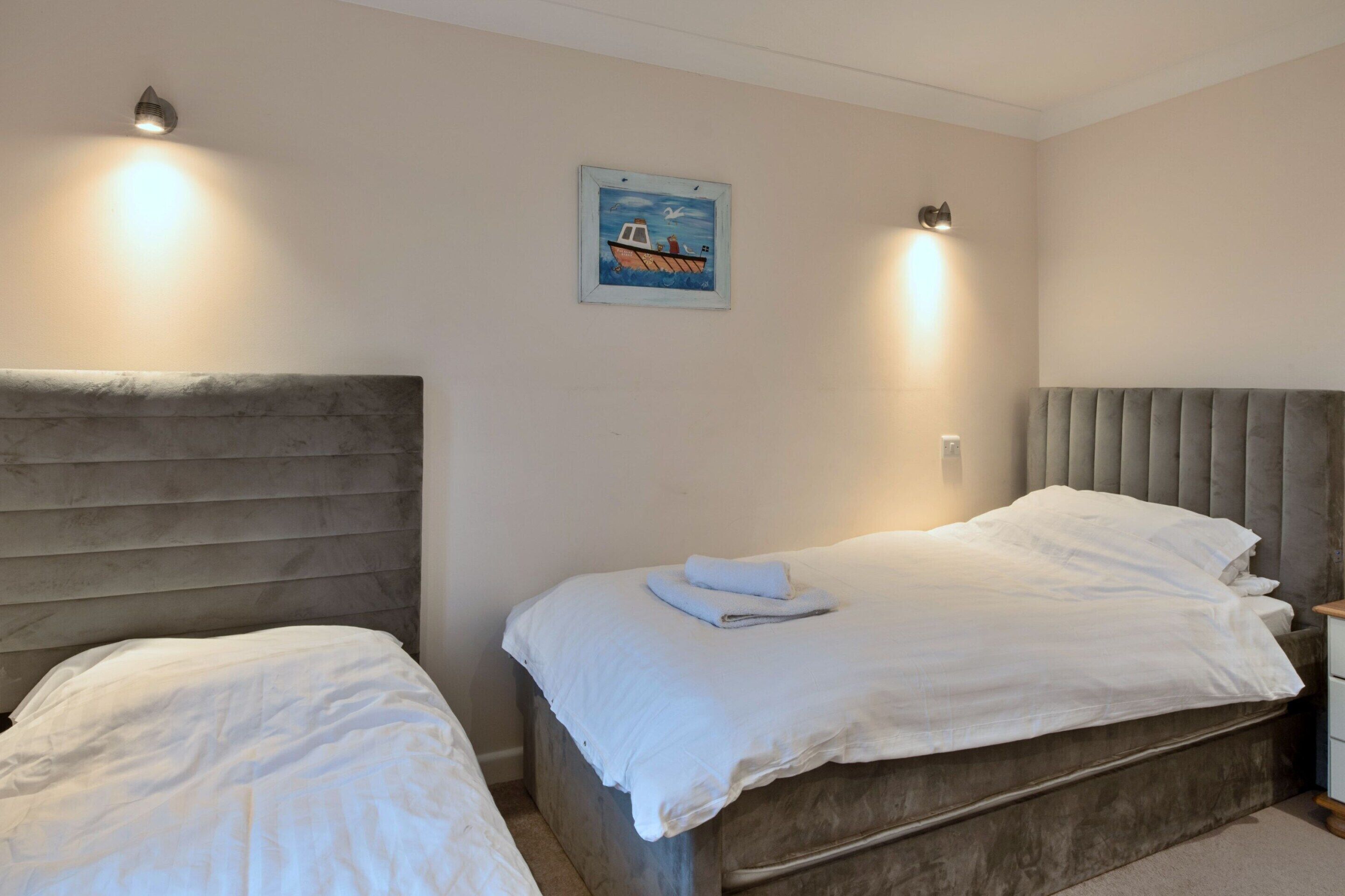 A bedroom with two beds, a headboard, a nightstand, a wall-mounted light, and a framed picture.