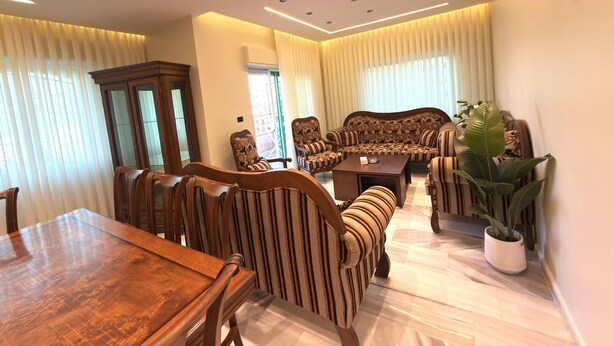 A spacious living area with a wooden dining table, striped upholstered chairs, a patterned sofa, and a potted plant. - Luxury 3-Bedroom Family Apartment, Prime Location, Free WiFi & Free Parking (Amman)