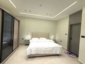 A modern bedroom with a large bed, two bedside tables with lamps, a sliding glass door, and a wooden door. - Luxury 3-Bedroom Family Apartment, Prime Location, Free WiFi & Free Parking (Amman)