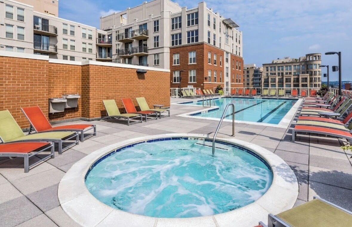 A rooftop pool area with lounge chairs and a hot tub, surrounded by apartment buildings.