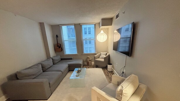 Ein Wohnzimmer mit einem Ecksofa, einem kleinen weißen Sofa, einem Couchtisch, einem an der Wand montierten Fernseher und einer Gitarre, die an der Wand hängt.