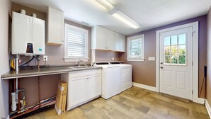 Une cuisine avec des meubles blancs, un four encastré, un lave-vaisselle et une fenêtre avec des stores.