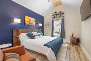 A bedroom with a bed, bedside lamps, a painting, a wooden dresser, and a window with curtains. - Mid-Century Modern Retreat with Hot Tub (Branson)