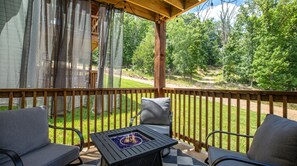 A covered outdoor seating area with a fire feature, wooden deck, and a view of a forested area. - Mid-Century Modern Retreat with Hot Tub (Branson)