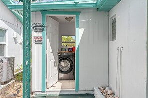 Un cuarto de lavandería con lavadora, un cartel que prohíbe fumar y vapear, y un pequeño estante con productos de limpieza.