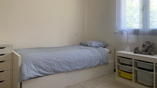 Een kinderkamer met een bed, een raam met gordijnen, een bureau met opbergmanden en een nachtkastje met een lamp.
