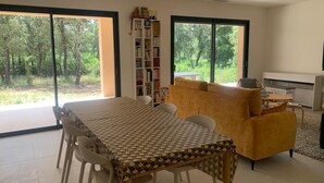 Een moderne woonkamer met een eettafel, bank en een boekenkast. Grote ramen bieden uitzicht op een bebost gebied.