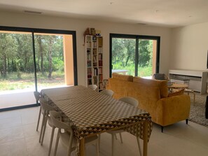 Ein modernes Wohnzimmer mit Esstisch, Sofa und Bücherregal. Große Fenster bieten Blick auf ein bewaldetes Gebiet.