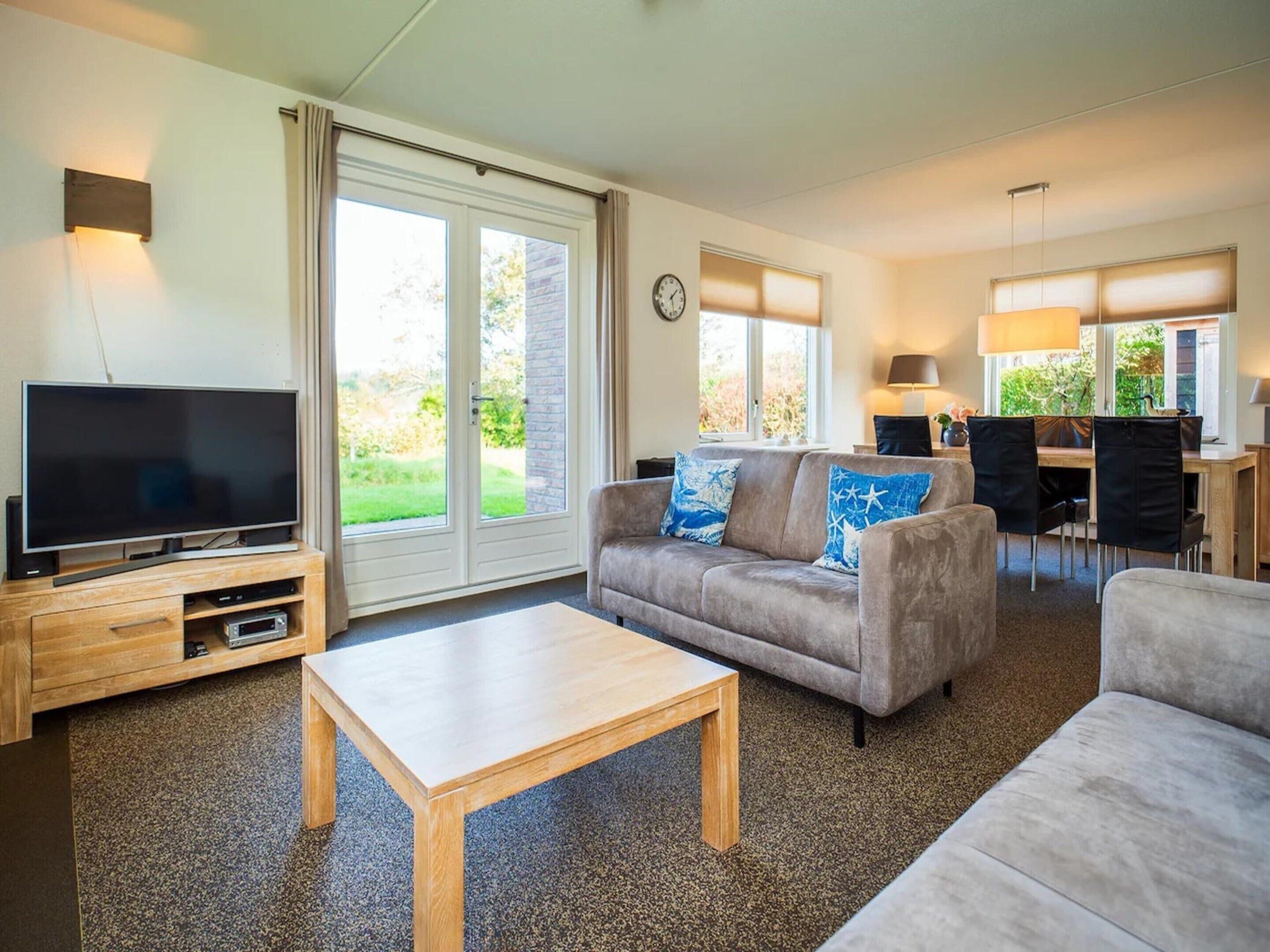 A living room with a flat-screen TV, a sofa with blue cushions, a wooden coffee table, and a dining area with chairs and a lamp.