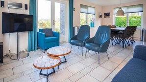 A modern living room with a flat-screen TV, blue armchairs, a dining table with chairs, and a large window with a view. - 3-bed Villa With Whirlpool (De Koog)