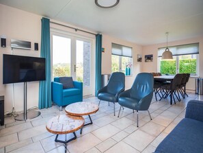 Ein modernes Wohnzimmer mit Fernseher, blauen Sesseln, einem Esstisch mit Stühlen und einem großen Fenster mit Blick.