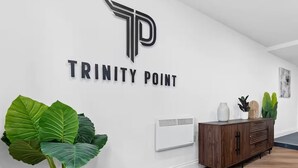 A hallway with a white wall, a large 'TRINITY POINT' sign, a wooden cabinet, and large green plants. - Stunning Spacious 2-bedroom apartment near London (Gravesend)