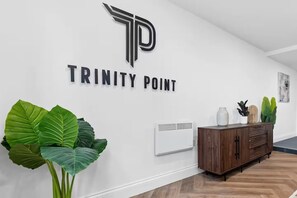Ein Flur mit weißer Wand, einem großen 'TRINITY POINT' Schild, einem Holzschrank und großen Grünpflanzen.