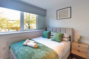 A bedroom with a bed, a window with a view of trees, a bedside table with a lamp, and a framed picture on the wall. - Stunning Spacious 2-bedroom apartment near London (Gravesend)