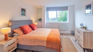 A modern bedroom with a bed, bedside tables, a dresser, a window with a view, and a ceiling light fixture. - Stunning Spacious 2-bedroom apartment near London (Gravesend)