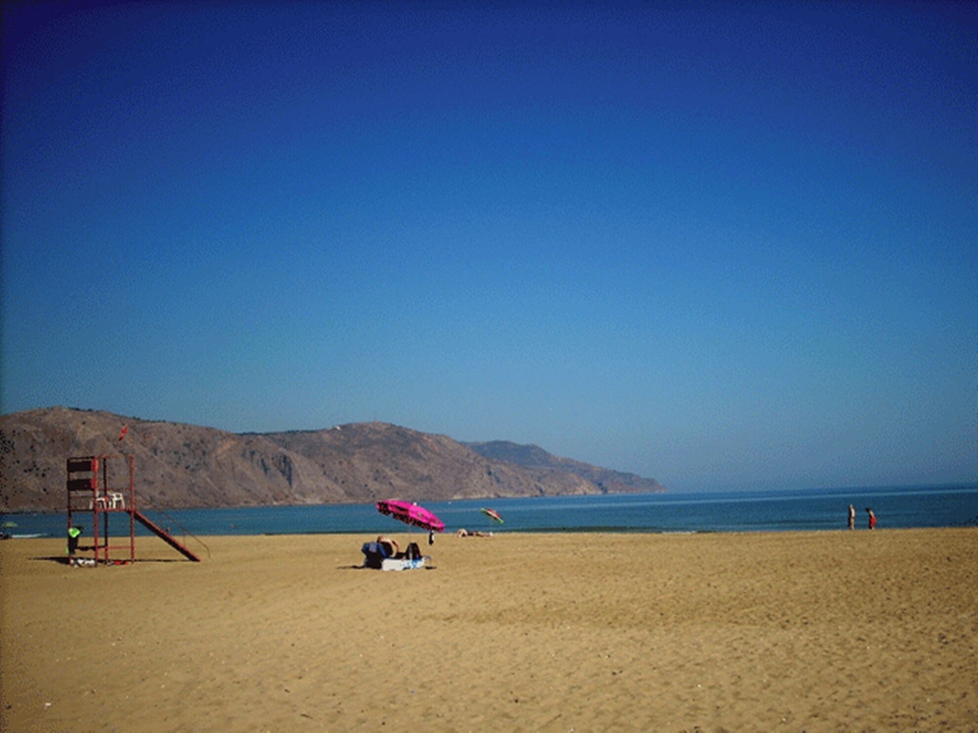 A beach with a lifeguard tower, a clear blue sky, and a distant mountain range.