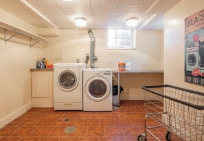 A laundry room with two front-loading washing machines, a sink, a window, and a poster on the wall. - Joseph Story Estate I Sleeps 12 l Parking + Elevator l On Salem Common (Salem)