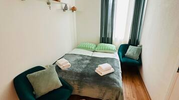 A small, cozy room with a bed, two chairs, a fur rug, and a shelf with decorative items.