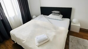 Una cama bien hecha con una manta y almohadas de punto blanco, una mesita de noche con una lámpara, y una ventana con cortinas.