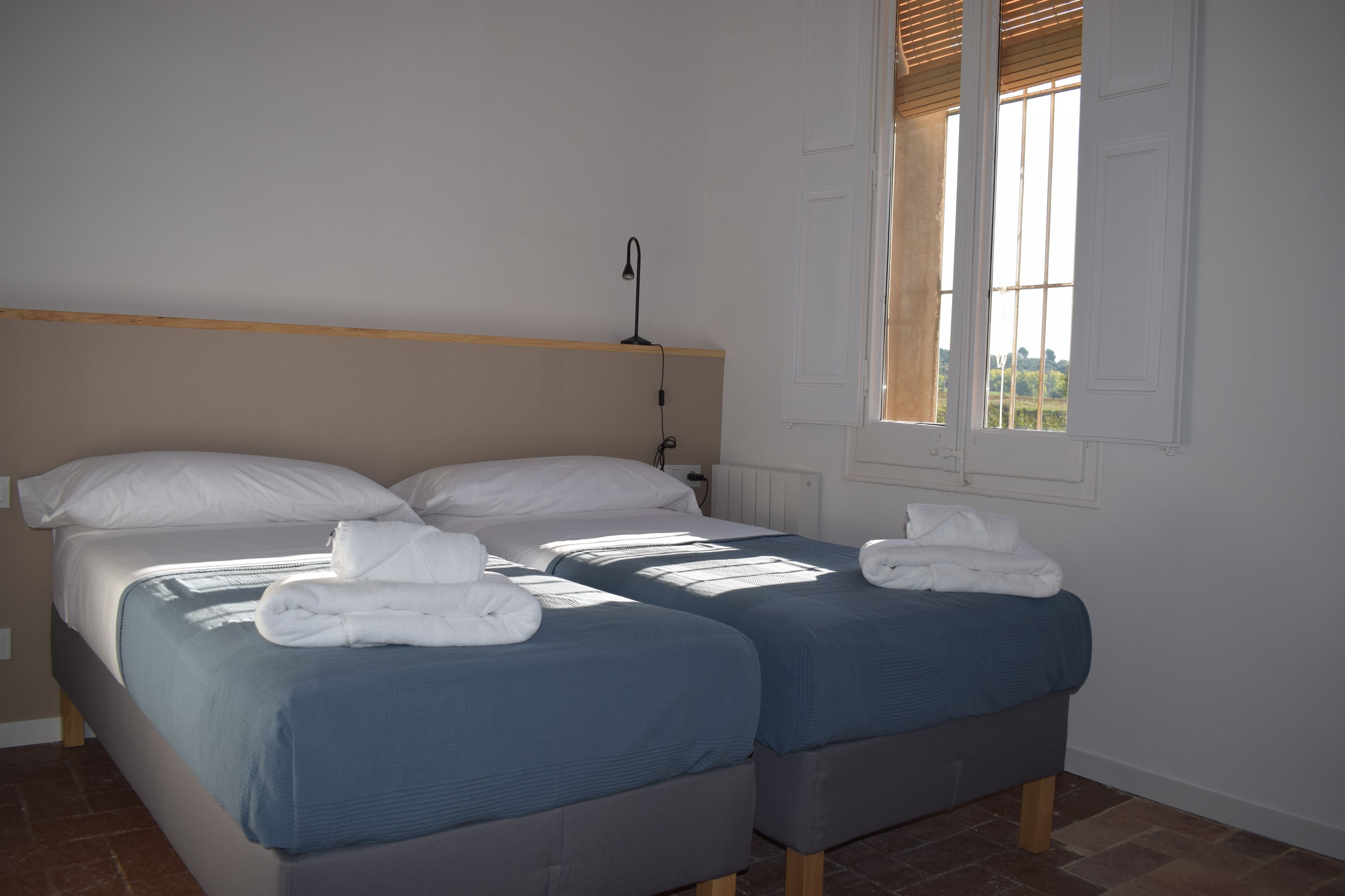 A room with two beds, each with a blue bedspread and white pillows, and a window with blinds.