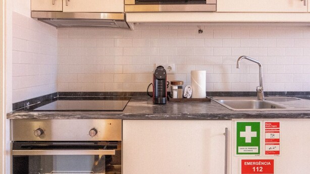 Eine Küche mit Mikrowelle, Backofen, Kaffeemaschine und Spüle. Notausgangsschilder und eine Erste-Hilfe-Ausrüstung sind sichtbar.