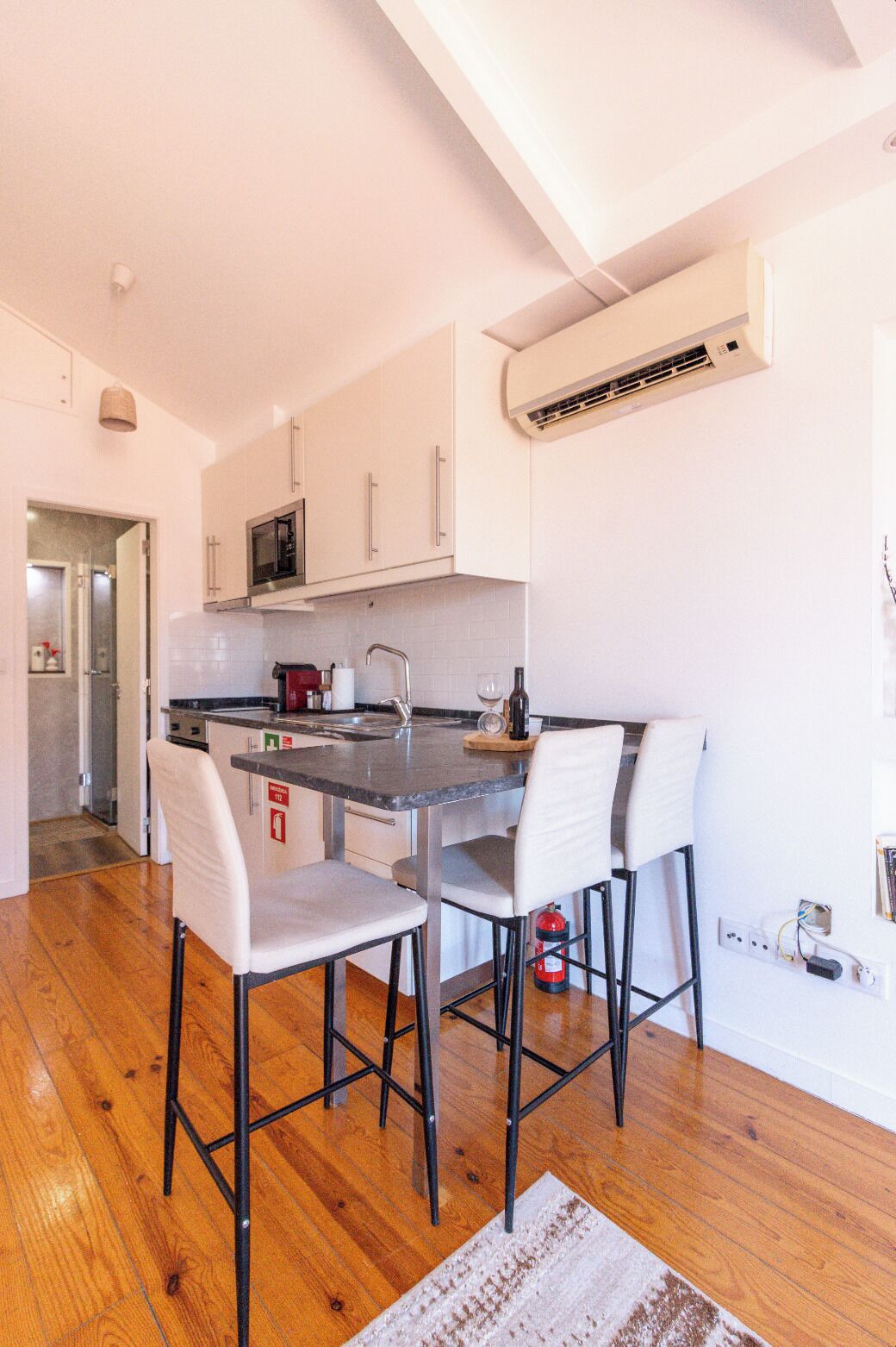 A modern kitchen with white cabinets, a microwave, and a built-in oven. There is a dining area with a bar stool and a small table.