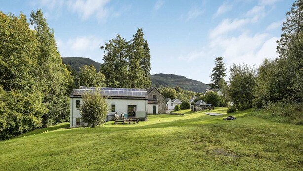 A house with solar panels, surrounded by a well-maintained lawn and trees, with a mountain in the background. - 9-bedroom Scottish Retreat (Colintraive)