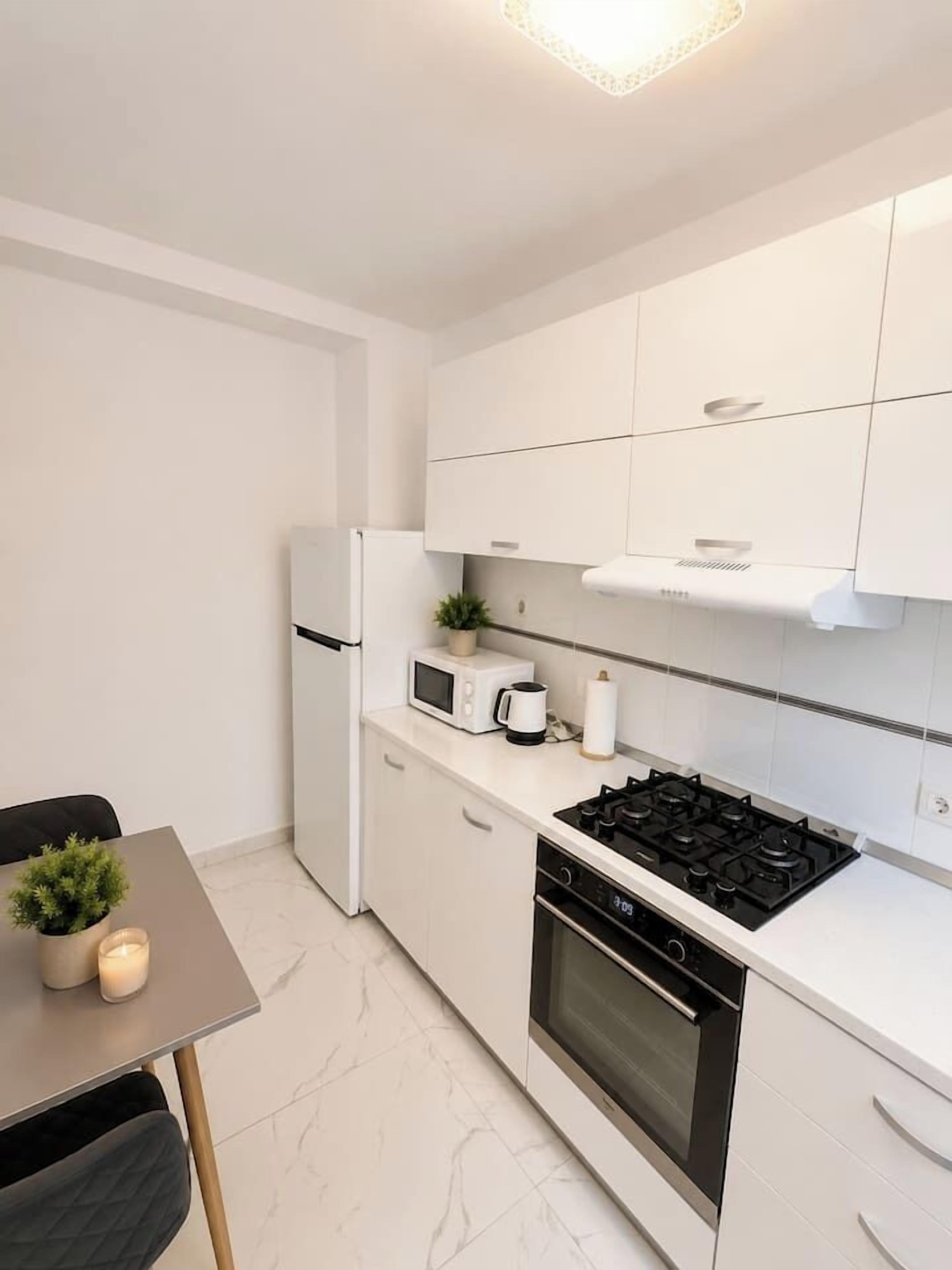 Une cuisine compacte avec des meubles blancs, une cuisinière, un four, un four à micro-ondes et une petite table à manger agrémentée d’une plante.