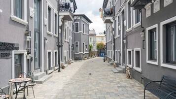 Une rue étroite bordée d’immeubles gris, avec un banc et des tables installées à l’extérieur.