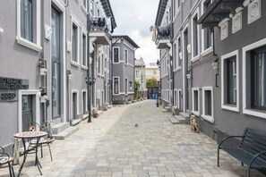 Eine schmale Straße mit grauen Gebäuden, einer Bank und Außenplätzen.