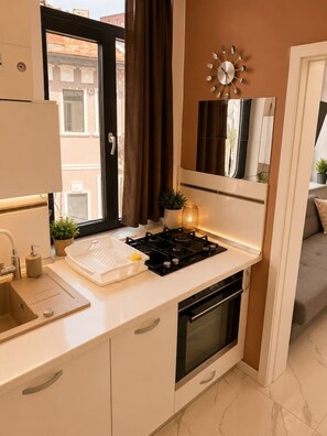 A modern kitchen with a gas stove, sink, and oven, a window with curtains, and a wall-mounted clock. - Stylish 1-Bedroom Apartment with Warm Accents (București)