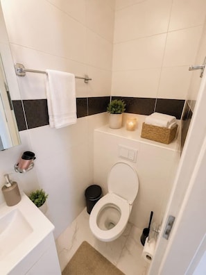 A compact bathroom with a white toilet, a small trash bin, a towel rack, a sink, and a potted plant. - Stylish 1-Bedroom Apartment with Warm Accents (București)