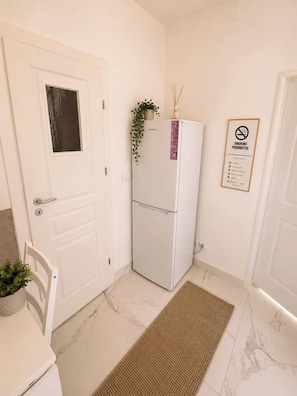 Een compacte keuken met een witte koelkast, een klein tafeltje met een plant, een deur met raam en een rookvrij bord aan de muur.