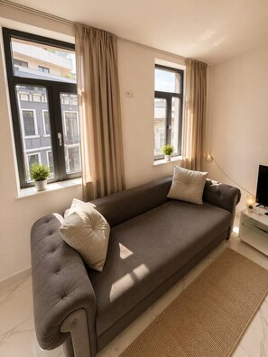 Ein modernes Wohnzimmer mit einer Couch, zwei Vorhängen an Fenstern, einer kleinen Pflanze und einem Fernseher auf einem Schrank.