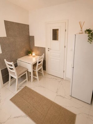 Eine kleine Küche mit einem weißen Tisch, zwei Stühlen, einem weißen Kühlschrank und einer Tür mit kleinem Fenster.