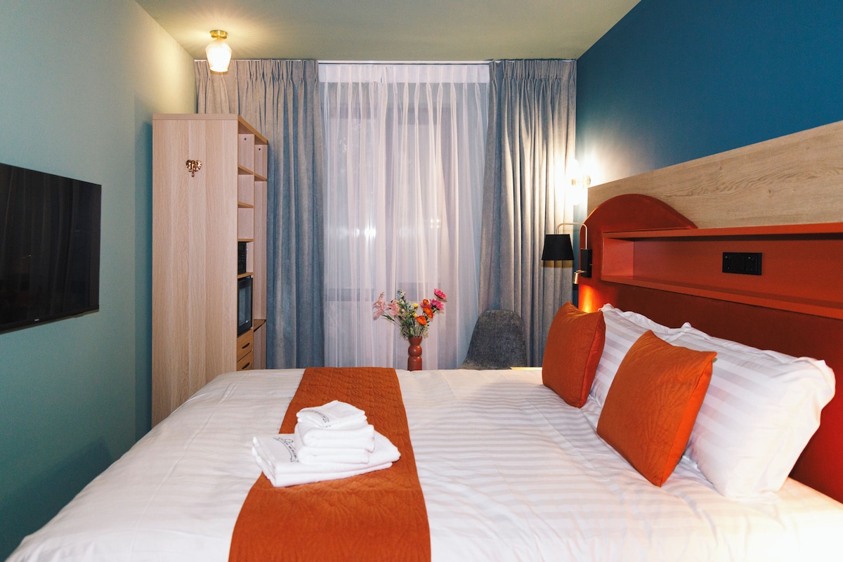 A hotel room with a bed, a wardrobe, a desk, a chair, a window with curtains, and a vase with flowers.