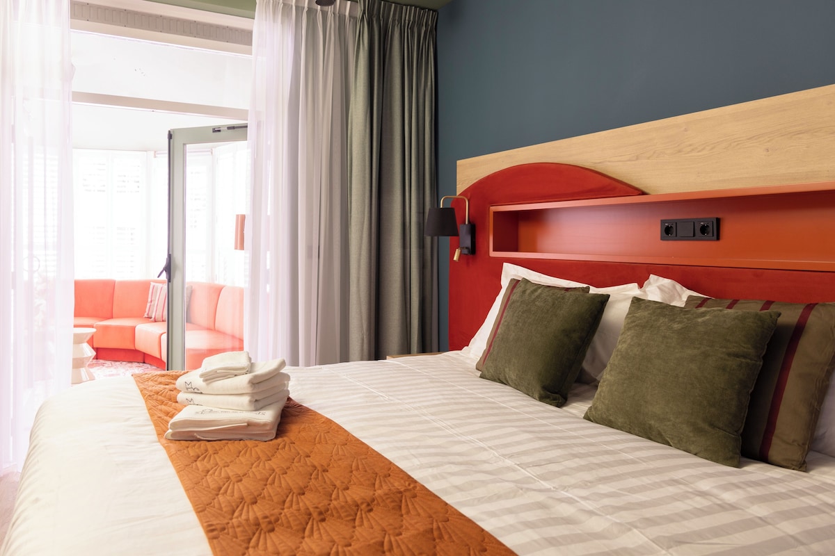 A hotel room with a bed, a red headboard, a window with curtains, and a view of the outside.